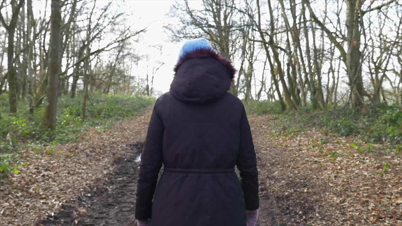 A SLOW MOTION GIMBAL SHOT following a woman from behind, walking through the woods on a footpath.