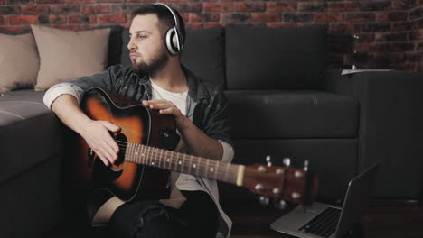 joven músico con guitarra, escuchando música con auriculares inalámbricos y usando una laptop en casa