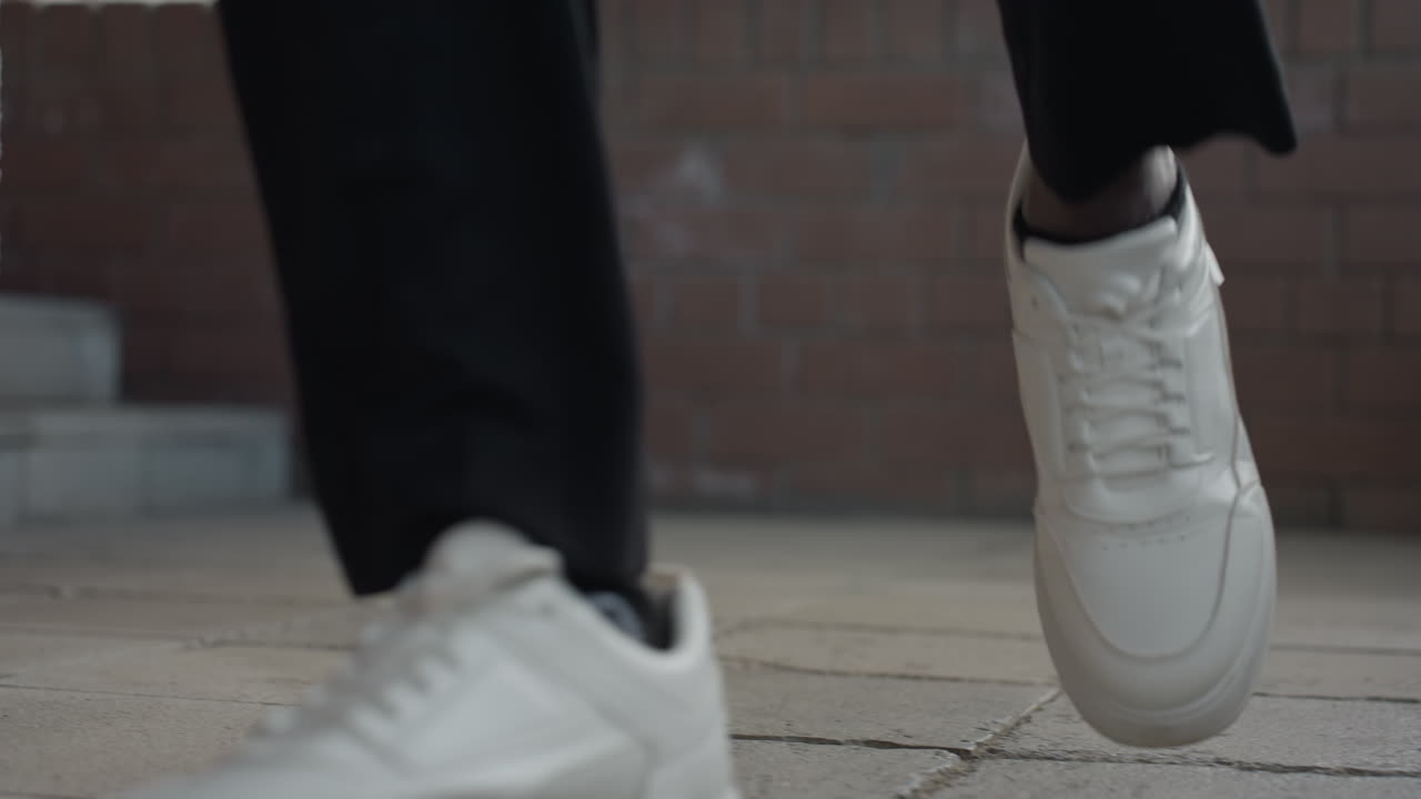 돌계단을 내려가는 흰색 스니커즈를 클로즈업한 사진이에요. 발 하나는 공중에 있고, 캐주얼한 바지가 보입니다. 은은한 빛이 도는 도시 입구에서 서두르는 듯한 움직임이 느껴지고, 깨끗한 신발은 질감이 있는 계단과 대비를 이룹니다.