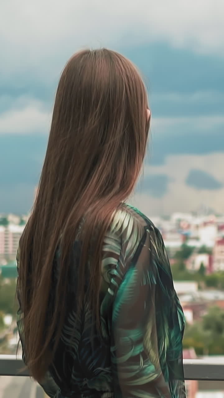 Stylish woman with long loose brown hair in dress with colorful decor looks at modern city from viewing deck slow motion close backside view