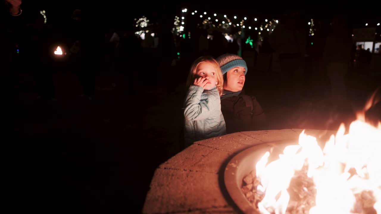 Mother and child at a firepit in winter with Christmas lights in the background at nighttime