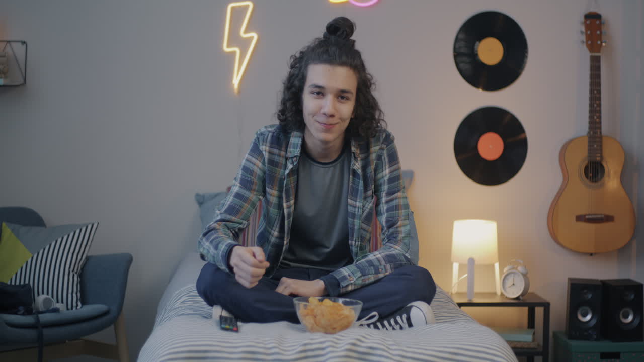 Teenage Boy Eating Chips and Watching TV in His Bedroom