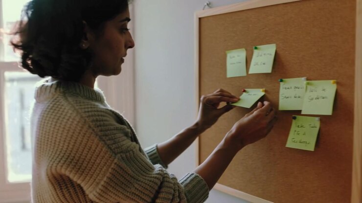 Closing laptop woman in sweater going to corkboard at home peeling sorting green notes for planning