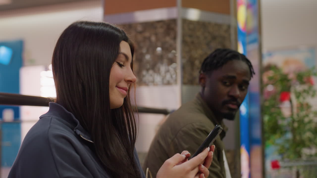 Woman smiling at phone while man beside her looks away with sad thoughtful expression in blurred shopping mall setting filled with vibrant lights, glass, and plants