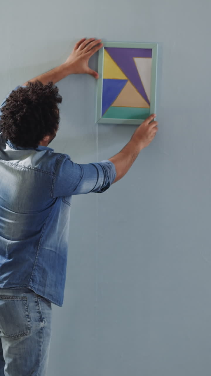 Black man helps colleague to hang drawing straight on wall in room backside view. Interior design practice and trends. Work upon domestic style