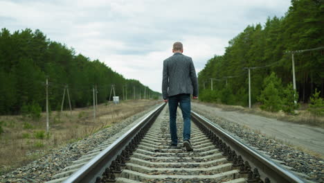 close view of a man wearing a suit jacket and jeans, walking alone on a railway tracks surrounded by trees, with electric poles around