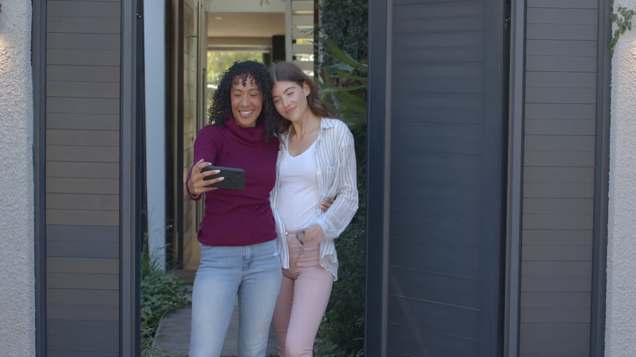 Taking selfie, lesbian couple smiling and standing together outside home