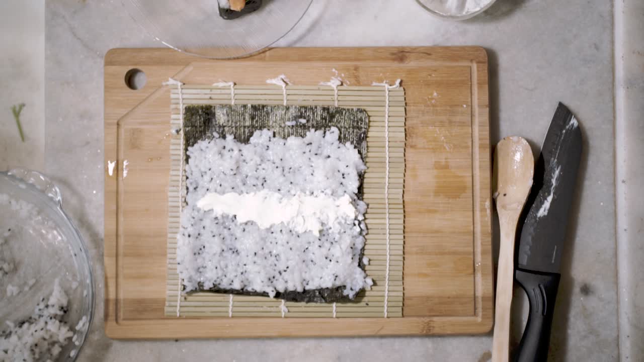 스시를 만들기 위해 매트에 차이 씨앗과 해초 노리 시트를 뿌린 부엌 카운터에 있는 나무 절단 태블릿에 두 손에 위에서 아래로 보기의 시간 경과