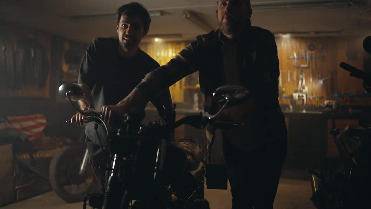 체크색 셔츠를 입은 자신감 있는 남성 기계공과 회색 티셔츠를 착용한 남성 고객이 수리 후 스튜디오 워크에서 오토바이를 꺼낸다.