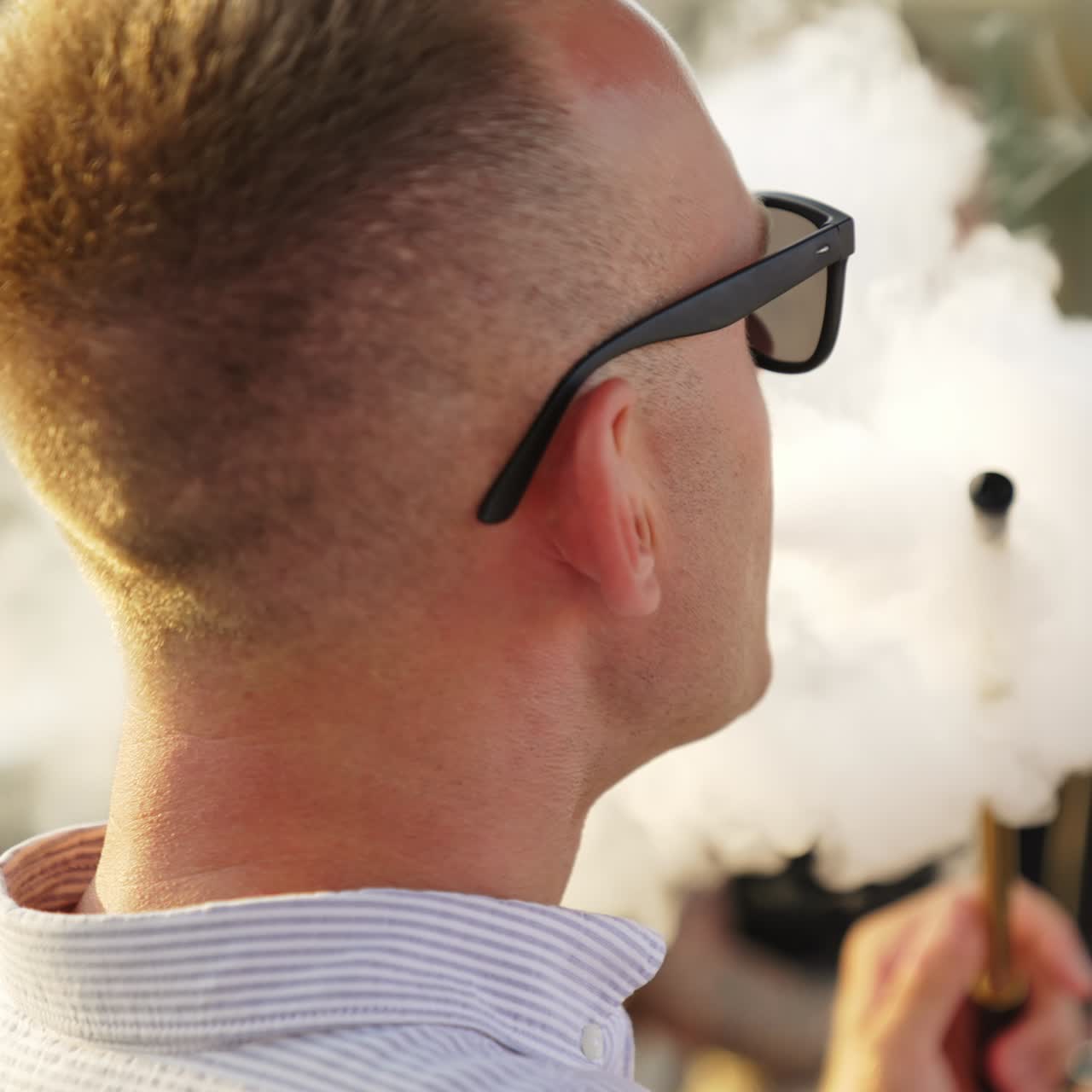 Calm Caucasian man wearing sunglasses smoking hookah sitting in a café outdoors. Girls sitting opposite at backdrop in blur