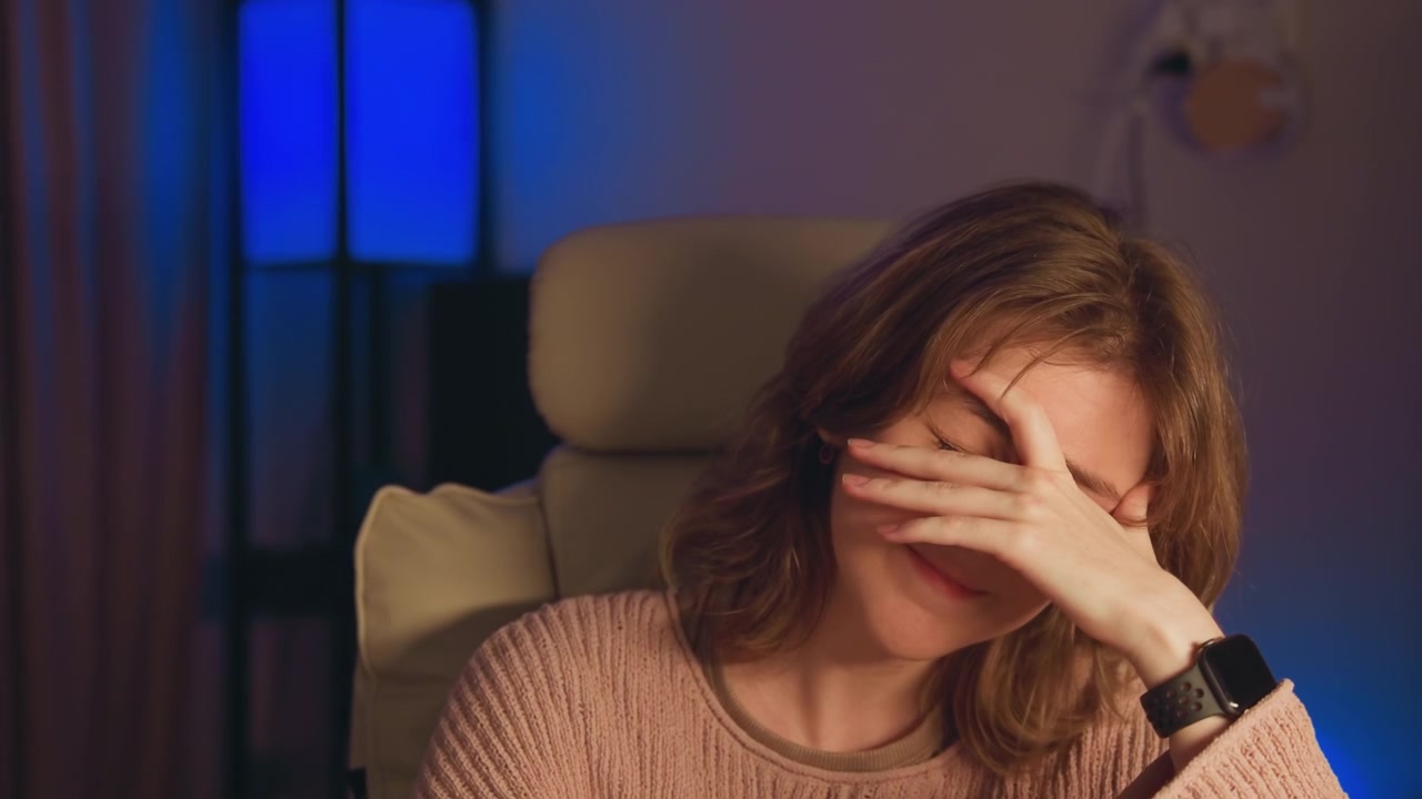 Ragazza adolescente seduta su una poltrona imbottita si copre il viso con la mano mentre sorride, mostrando divertimento timido, imbarazzo giocoso e un'atmosfera casalinga rilassata sotto una luce calda principale con un accento blu.
