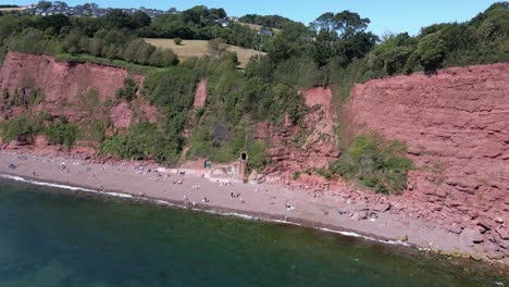 Toma-Aérea-Ascendente-Hacia-Atrás-De-Personas-Descansando-En-La-Playa-De-Ness-Cove-Con-Un-Acantilado-Rojo-En-El-Fondo-Y-El-Paisaje-Rural-De-La-Ciudad-En-La-Parte-Superior