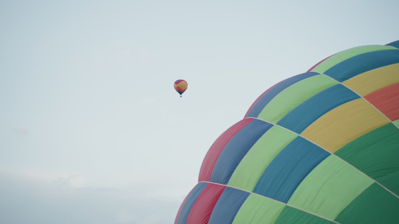 checker patterned hot air balloon envelope partially inflated in grassy field tethered by rope lines held by crew member while smaller balloon drifts overhead against pale sky