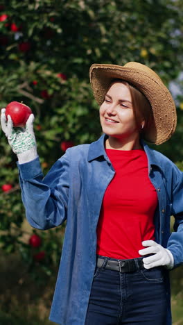sunny bright day on the harvest season in apple garden. happy smiling lady farmer holding an apple, looking at it and smelling it. vertical video