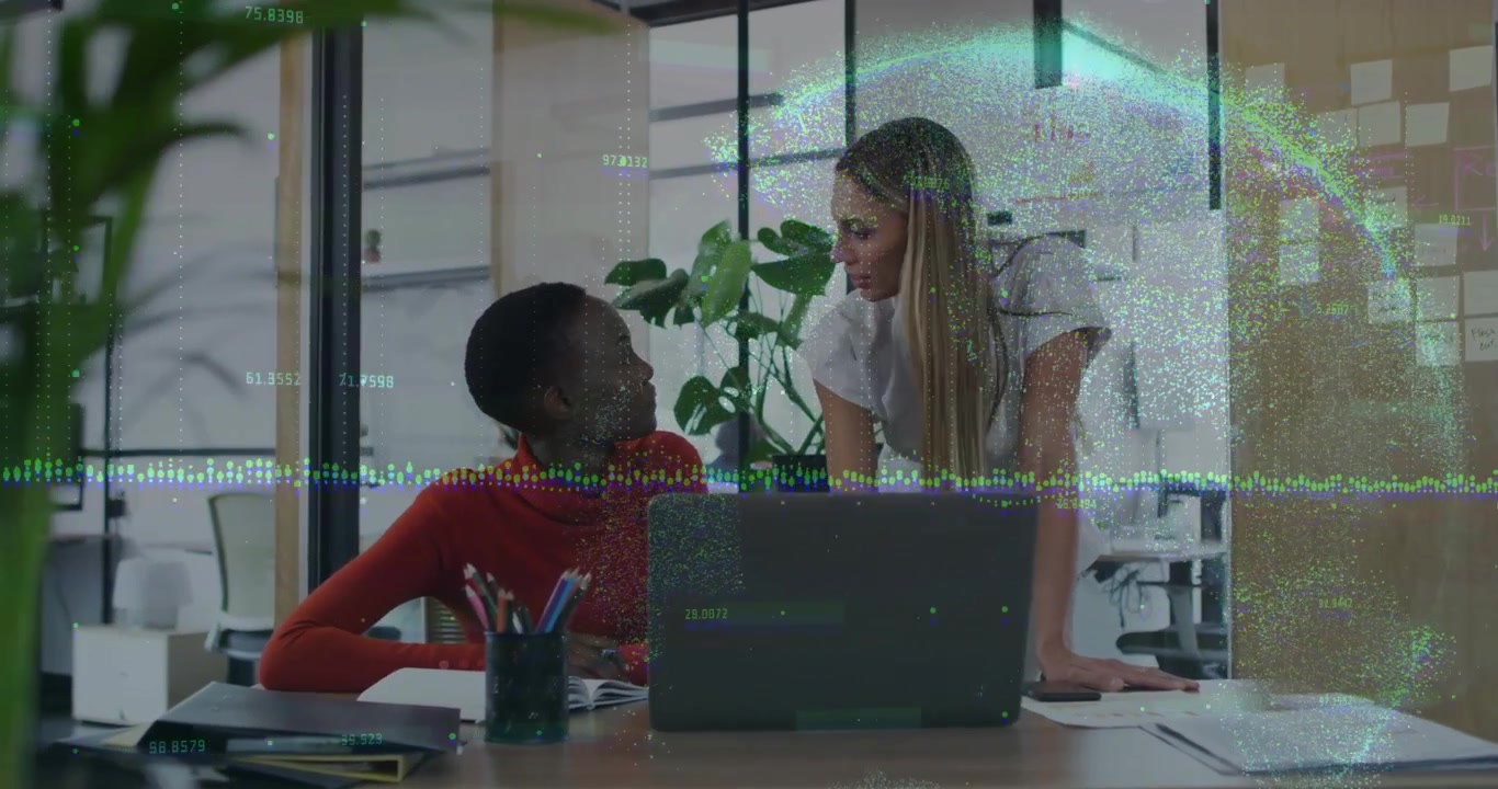 Seated woman taking business notes while colleague pointing at laptop starting graphs expanding