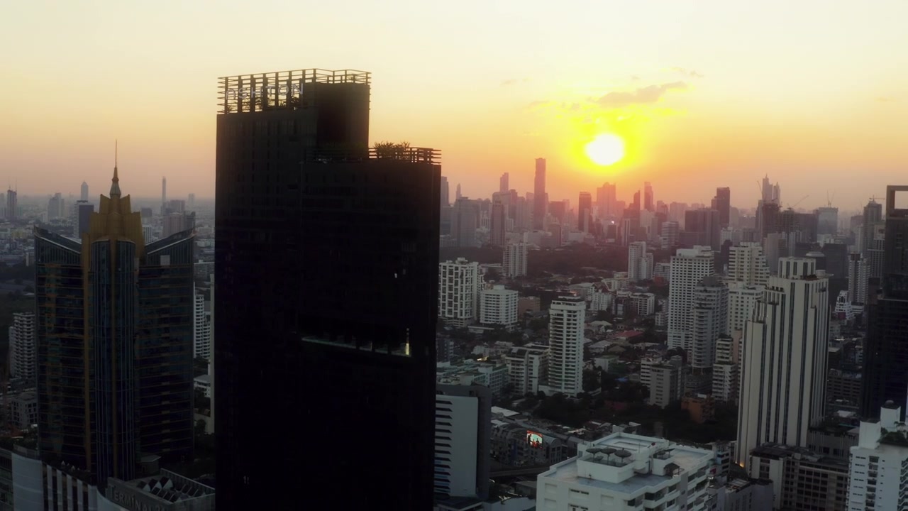 Um drone sobrevoa uma torre escura e um telhado dourado em direção ao horizonte de Bangkok ao nascer do sol.