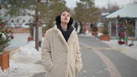 young lady in warm beige winter coat with black hoodie and earmuffs turns calmly, gazing toward bright sunlight. snowy street lined with decorated trees and festive atmosphere