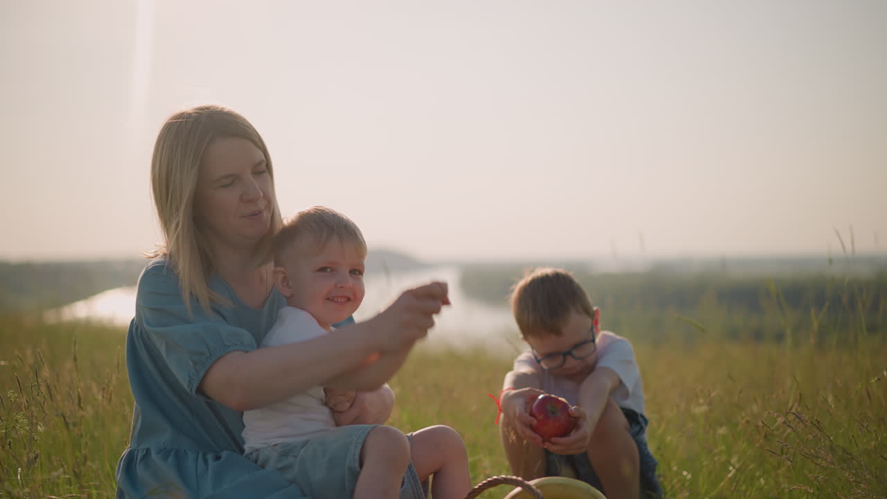 A cheerful mother in a blue dress enjoys a tender moment with her young son on her lap, gently holding his hands as they sit on the grass. In the background, the older brother is seen holding an apple