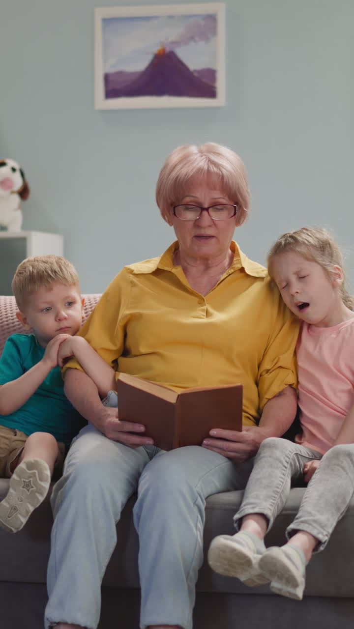 Careful mature woman reads fairy tales to sleepy little children on sofa at home. Loving grandmother spends evening with boy and girl. Babysitting process