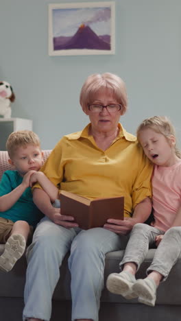 careful mature woman reads fairy tales to sleepy little children on sofa at home. loving grandmother spends evening with boy and girl. babysitting process