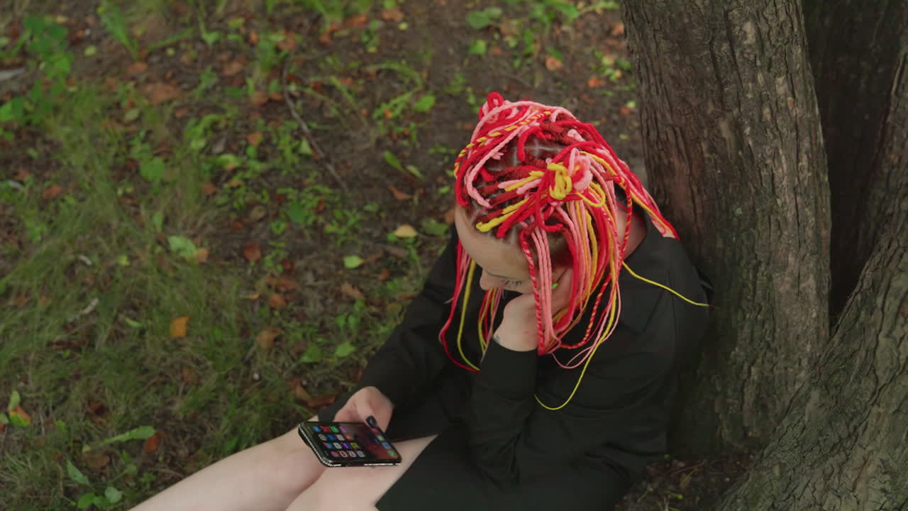 Plano general de una mujer caucásica con rastas de neón sentada bajo un árbol, mirando el teléfono con auriculares, hojas del parque alrededor de las piernas, abrigo de tono apagado que contrasta con el pelo vibrante, gesto de desplazamiento que transmite aislamiento