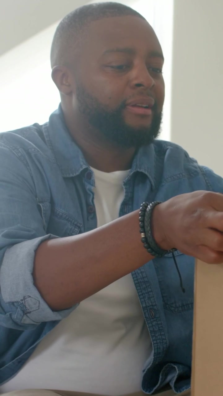 Vertical video: Box on lap, man in denim shirt, bracelets peeling tape and inspecting, living room