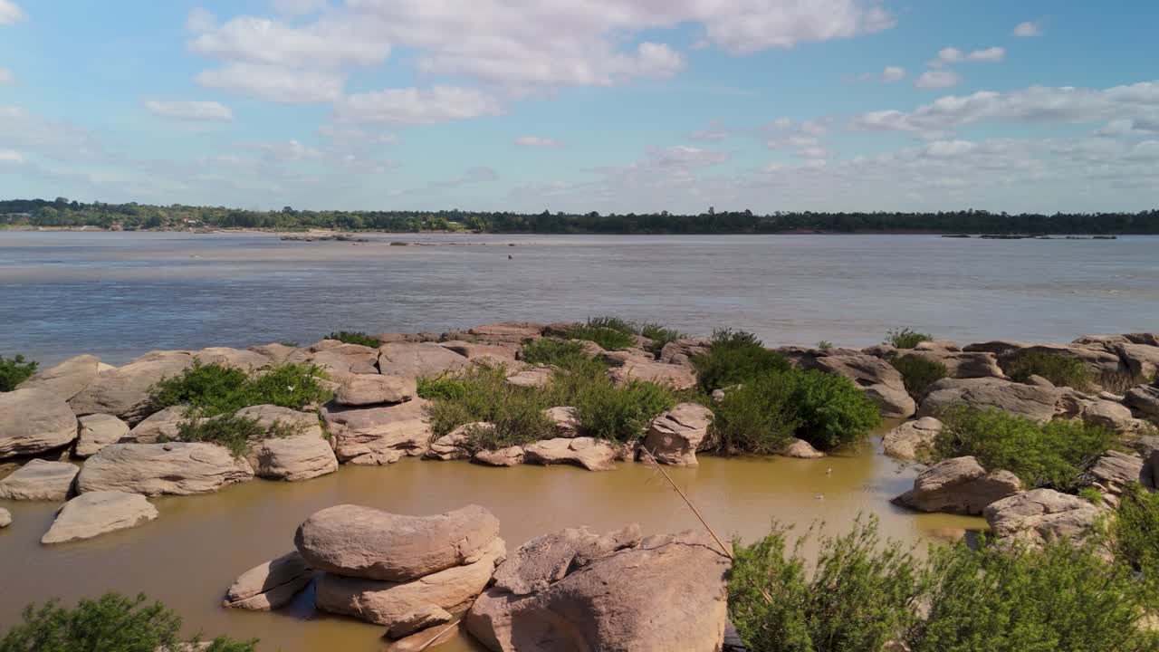 Alternate angle across the rocky Mekong shoreline with scattered bushes, soft clouds drifting over broad water