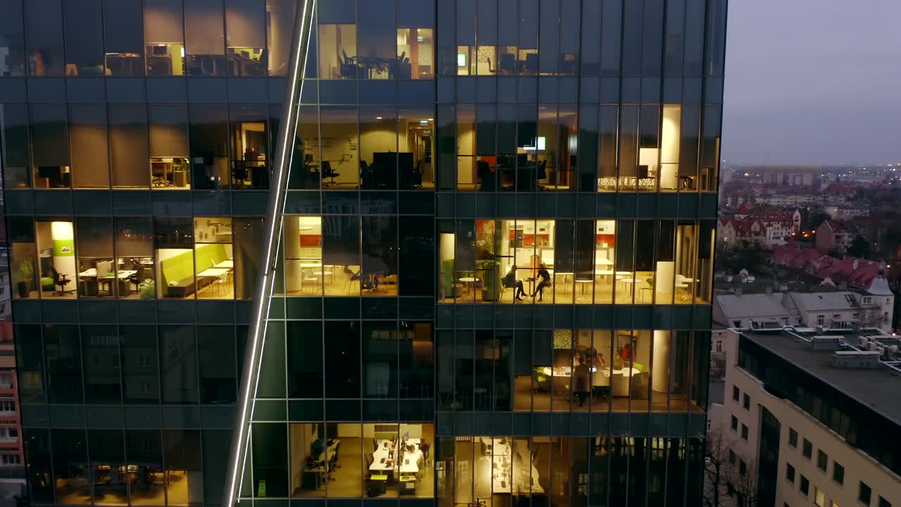 un dron disparó a un rascacielos nocturno con ventanas de oficinas y gente de negocios trabajando hasta tarde en las computadoras.