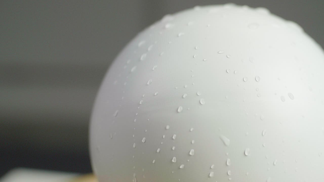 Close up of a white plain sphere wet spinning with reflecting water drops on the sphere. A dark gray background. Shot in Slow Motion in 4K.