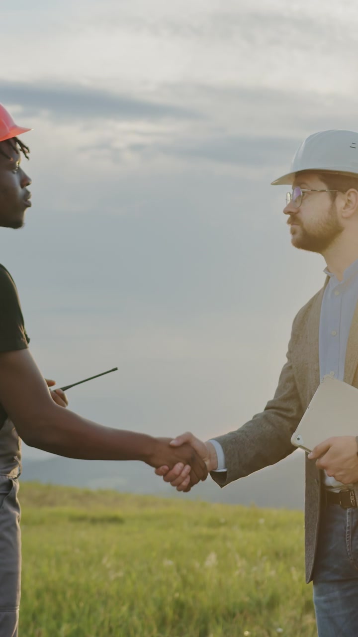 Due ingegneri con cappelli si stringono la mano in un campo con turbine eoliche sullo sfondo, discutendo di un