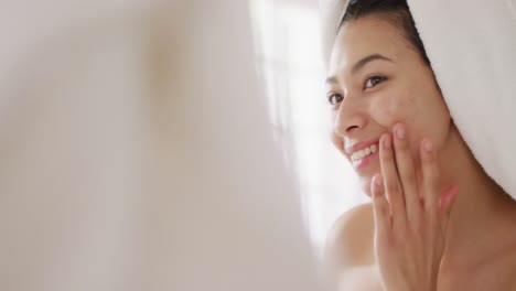 video of portrait of smiling biracial woman with towel on hair pplying cream in bathroom