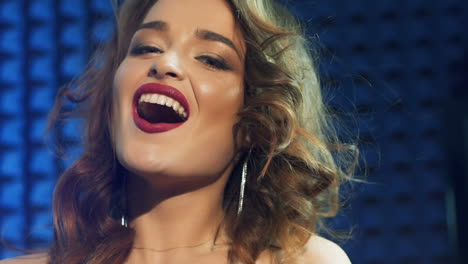 slow motion of gorgeous woman with bright make-up looking at camera and smiling isolated on blue background. portrait of a lovely singer with long wavy hair. close-up