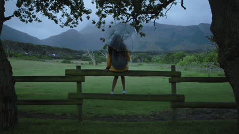 Mujer-Joven-Sosteniendo-Un-Paraguas-Sentada-Despreocupada-En-La-Valla-Disfrutando-De-La-Vista-De-La-Hermosa-Y-Tranquila-Granja-Campestre-Preparada-Para-El-Clima-Lluvioso