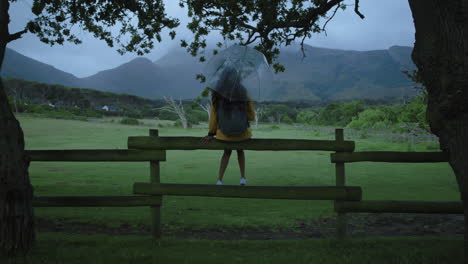 young woman holding umbrella sitting carefree on fence enjoying view of beautiful calm countryside farm prepared for rainy weather