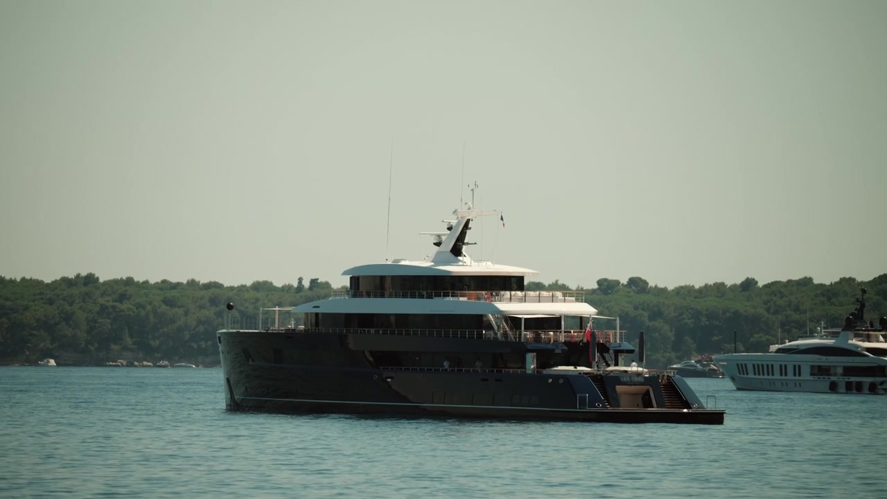 Superyacht che naviga in mare calmo, a simboleggiare ricchezza, tempo libero e uno stile di vita di viaggio di lusso. Alberi verdi che costeggiano lo sfondo costiero, Golfe Juan, Francia.