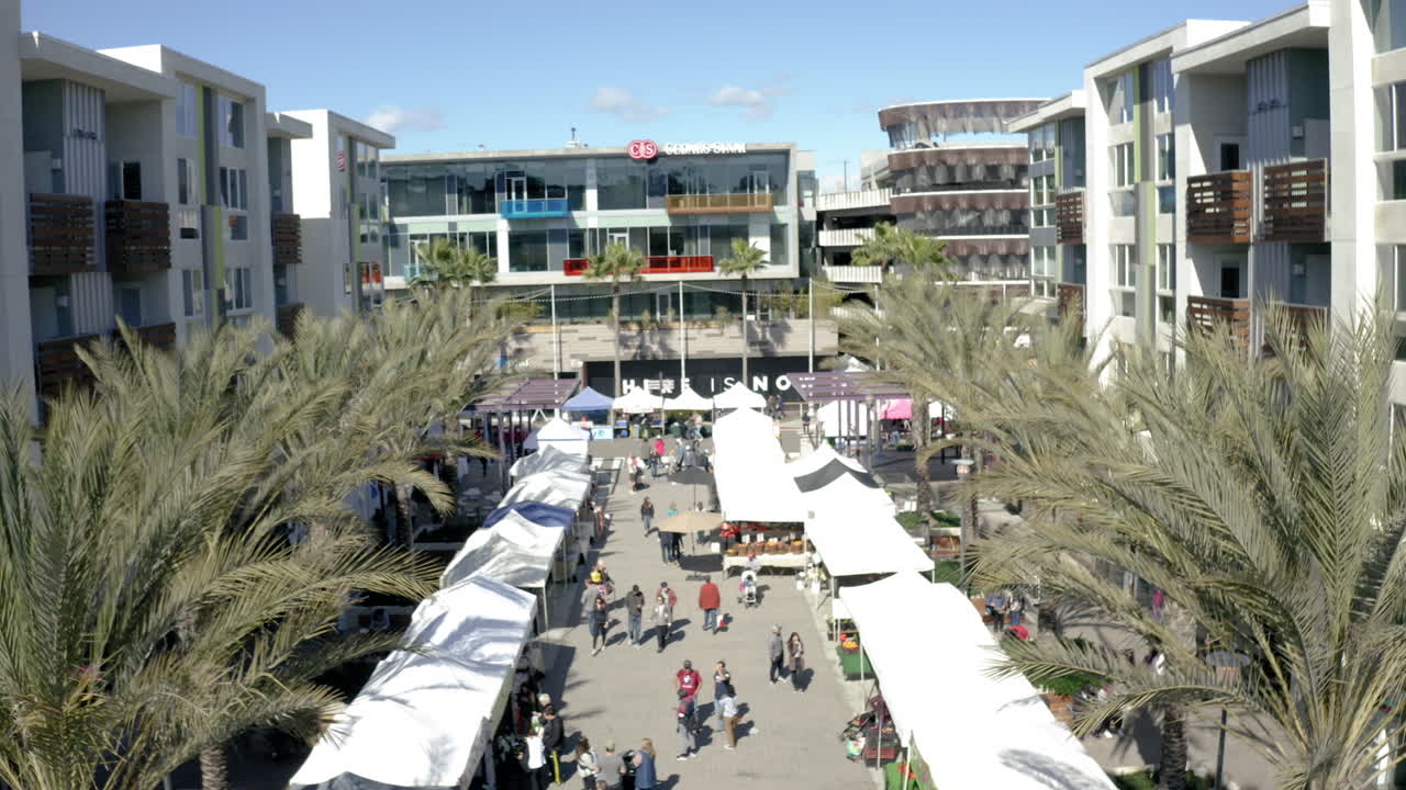 An outdoor market with numerous stalls and people walking between modern residential and commercial buildings on a sunny day