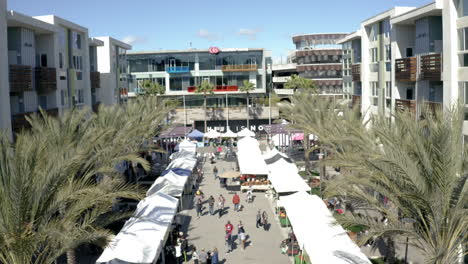 an outdoor market with numerous stalls and people walking between modern residential and commercial buildings on a sunny day