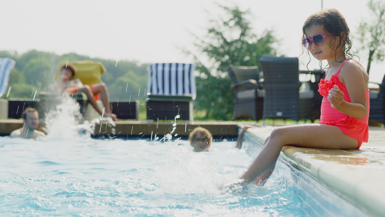 Pige sidder på kanten af udendørs swimmingpool og plasker fødder i vandet på familie sommerferie