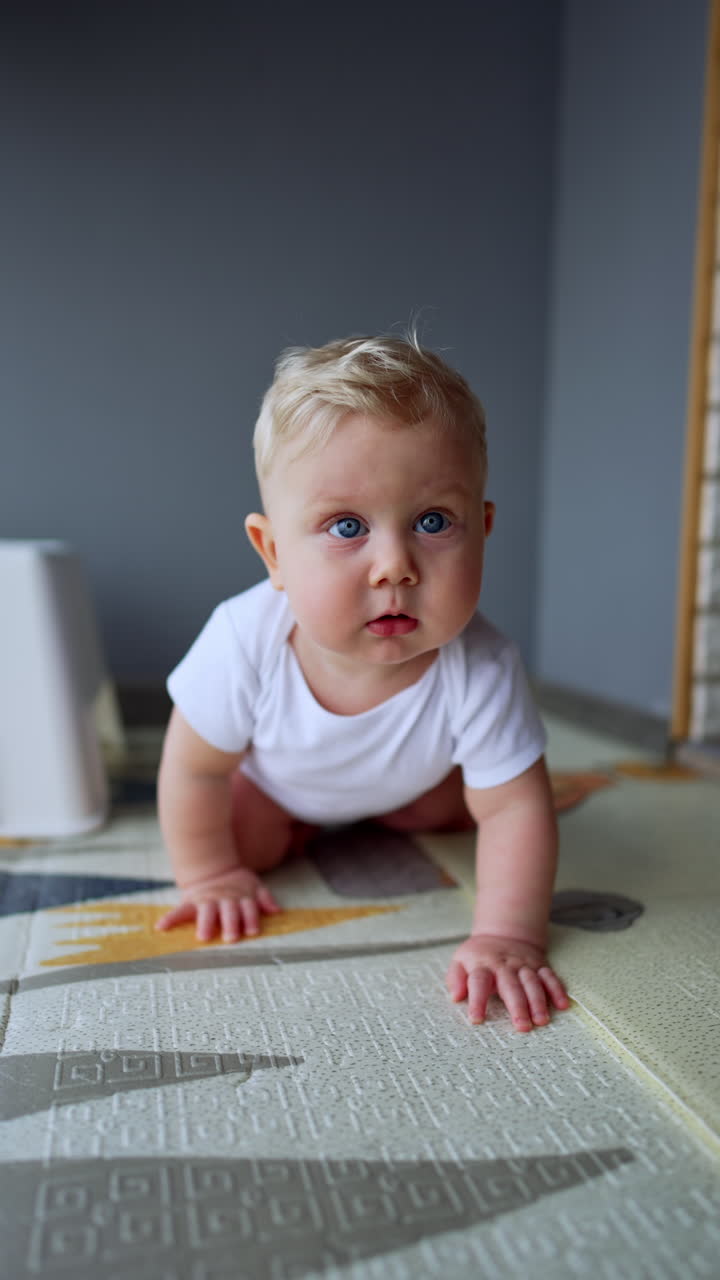 Chubby blond baby boy wearing white bodysuit tries to stand up. Child stands on all four and crawls to camera. Vertical video.