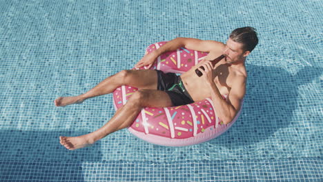 fotografía aérea de un hombre en pantalones cortos de natación flotando en un anillo inflable en una piscina bebiendo cerveza