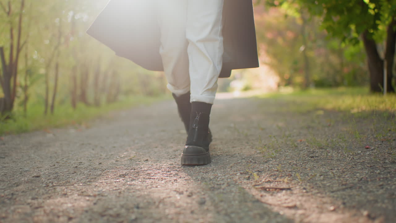 Low view of young girl in black coat and white trousers wearing boots, spinning and strolling along sunlit autumn park path, steps kicking gravel, playful motion, relaxed pace, soft breeze, greenery