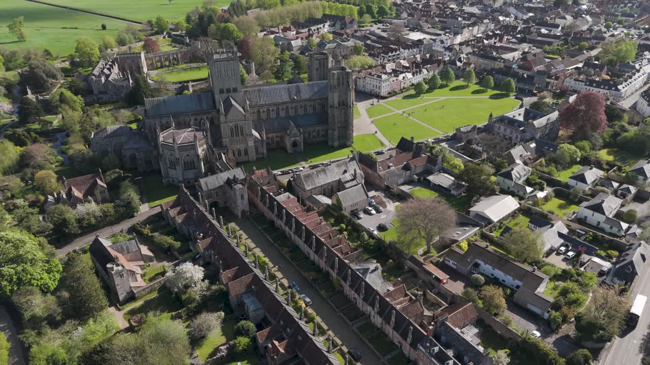 Orbital aerial of medieval Wells where soaring Gothic cathedral towers anchor compact historic townscape, blending heritage architecture, gardens and human-scale streets for cultural storytelling