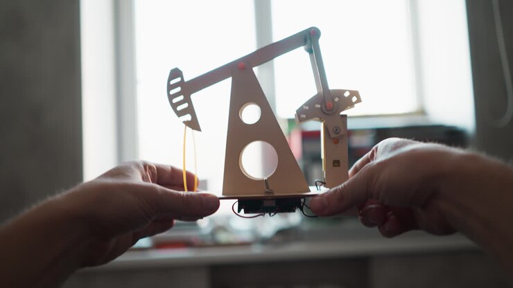 POV of person lifting a motorized oil pumpjack model to window level at a work desk, checking the electronics