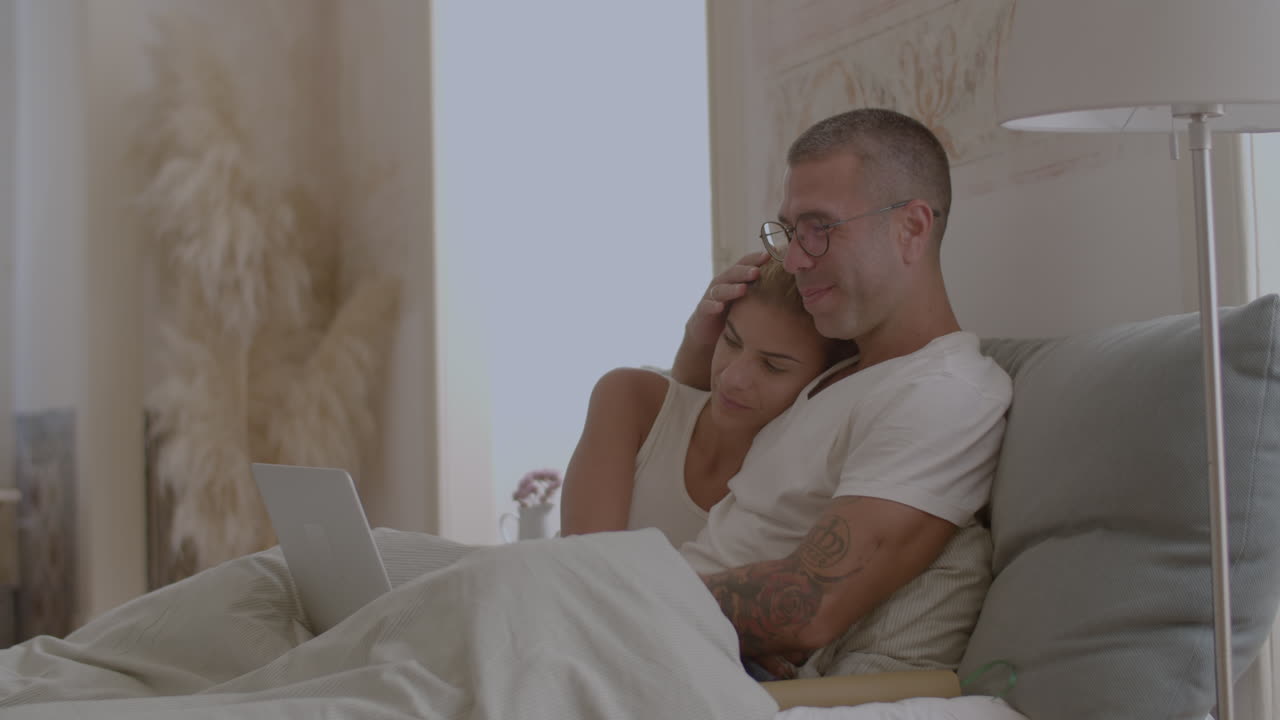 Happy man and woman sitting on bed and watching movie on laptop
