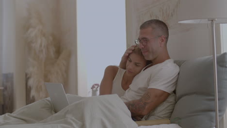 happy man and woman sitting on bed and watching movie on laptop