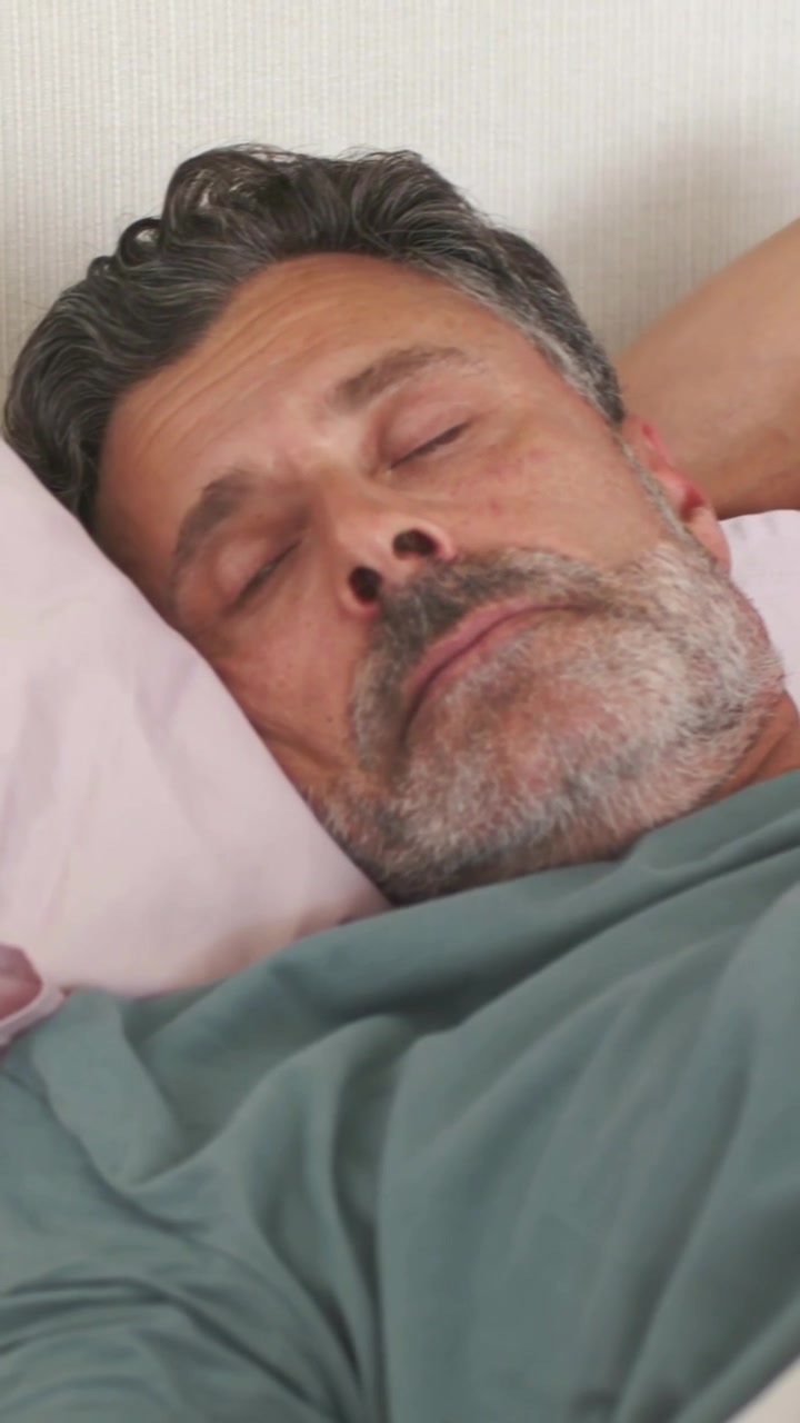 Video vertical: Hombre despertándose y abriendo los ojos en la cama, empujando una almohada rosa para acomodarse, con camiseta verde.