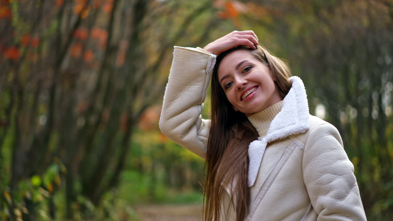 Happy resilient brunette lady smiling into camera. Woman in white jacket touching her hair and looking down shyly. Autumn park backdrop.