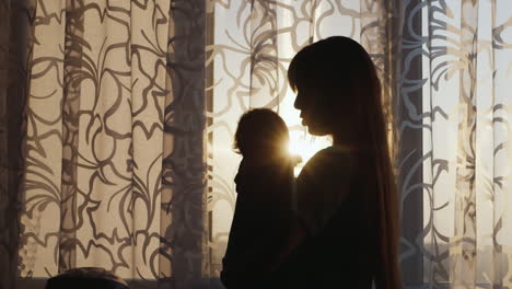 silhouette of a young mother with a child at the window swaying the baby in her arms at sunset