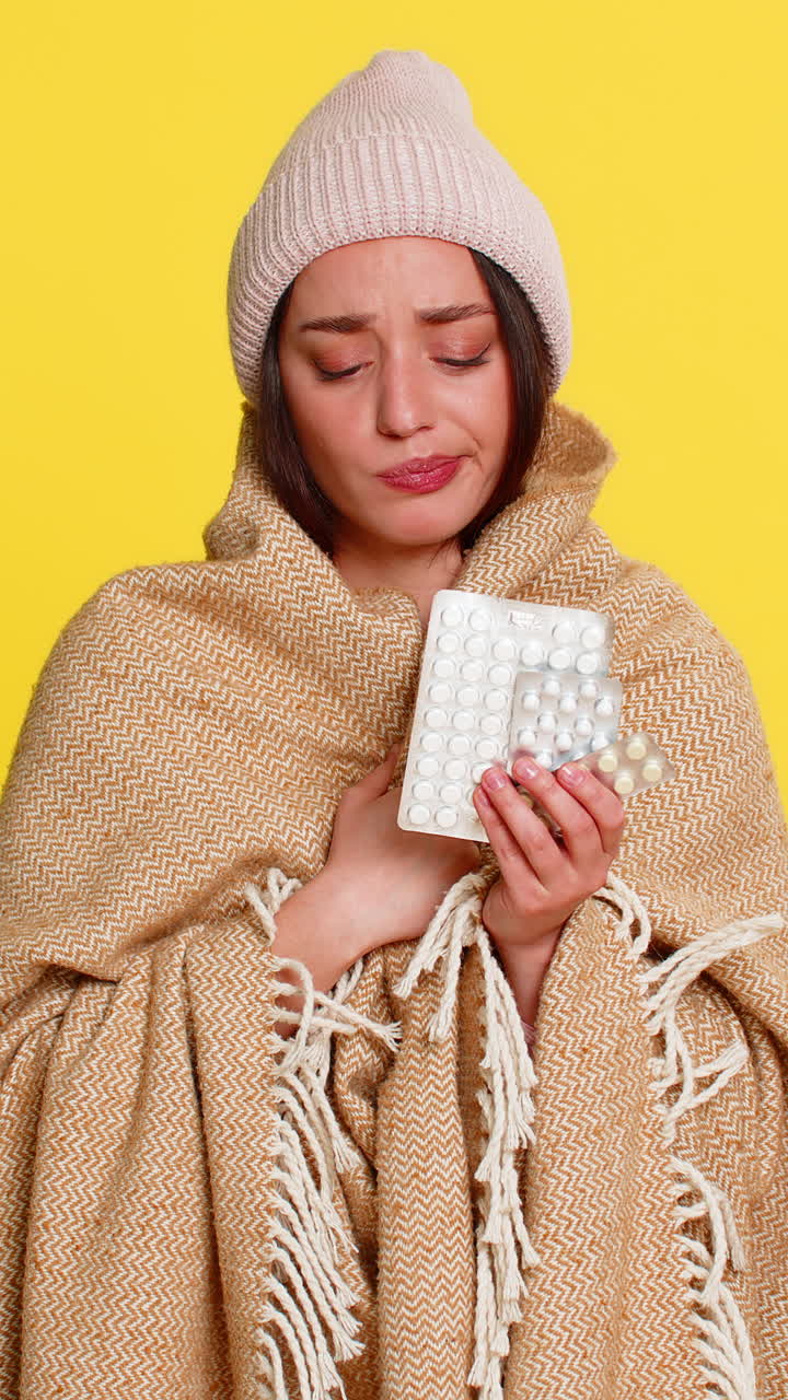 Donna avvolta in una coperta con un cappello che guarda delle pillole in modo pensieroso per curare un raffreddore o un'influenza