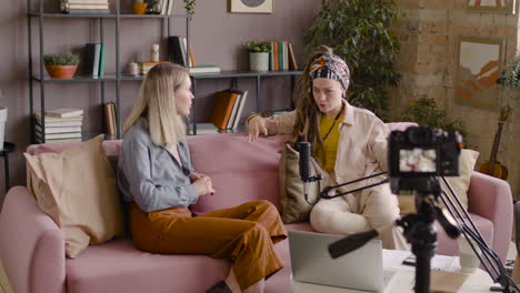 rear view of a camera thats is recording two women in a podcast talking into a microphone while they sitting on sofa